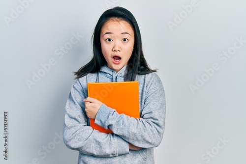 Young chinese girl holding book afraid and shocked with surprise and amazed expression, fear and excited face.