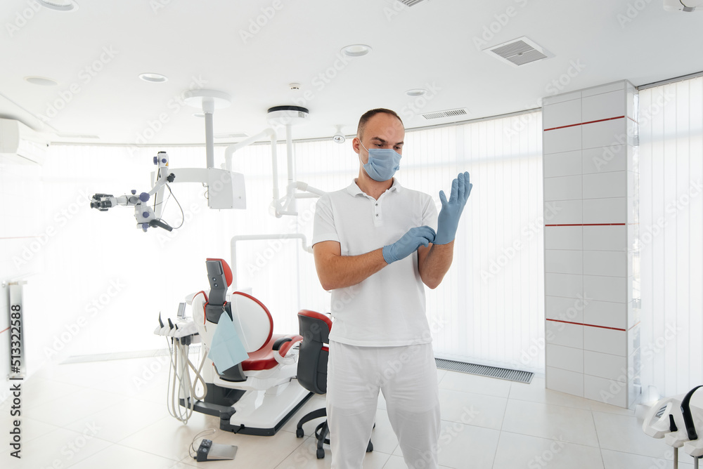 A young dentist in a mask stands near a red dental chair and smiles in modern white dentistry. Modern dentistry and prosthetics. A young dentist in a mask stands near a red dental chair and smiles in modern white dentistry. Modern dentistry and prosthetics.