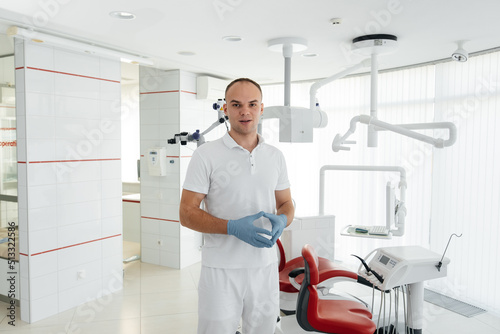 A young dentist stands near a red dental chair and smiles in modern white dentistry. Treatment and prevention of caries from youth. Modern dentistry and prosthetics.