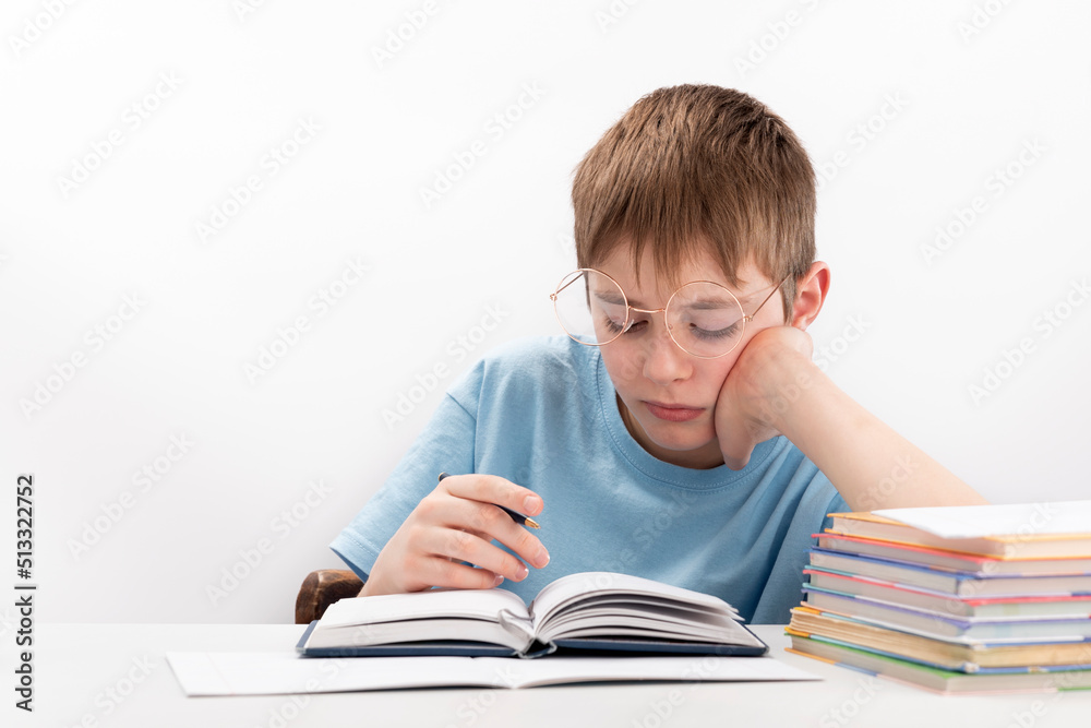 Bored boy doing homework at the desk. Schoolboy in glasses supports head with hand. Home schooling. Bored boy doing homework at the desk. Schoolboy in glasses supports head with hand. Home schooling.