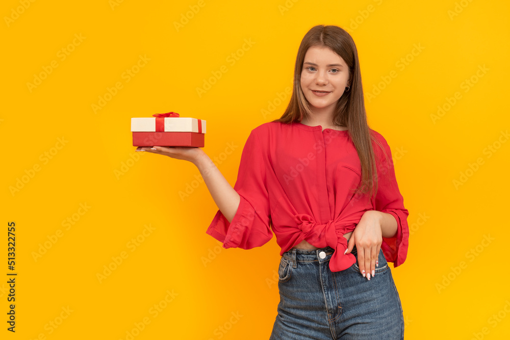 Smiling young girl holds gift box in palm of her hand on bright yellow background. Portrait of woman with gift Smiling young girl holds gift box in palm of her hand on bright yellow background. Portrait of woman with gift