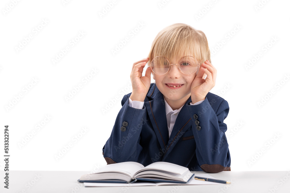 Happy funny blond boy in big glasses wears school uniform sitting at desk and smiling at the camera. Education concept. Isolated on white background. Happy funny blond boy in big glasses wears school uniform sitting at desk and smiling at the camera. Education concept. Isolated on white background.