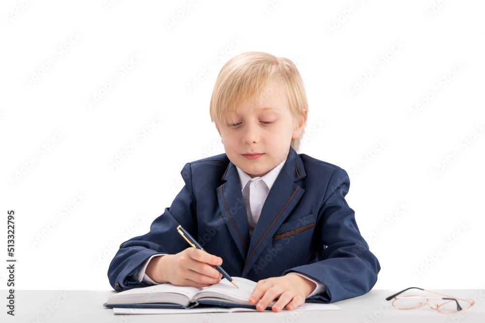 Diligent student writes in copybook. Child like boss sits at table and writes in notebook. Blond boy in jacket and shirt. Diligent student writes in copybook. Child like boss sits at table and writes in notebook. Blond boy in jacket and shirt.