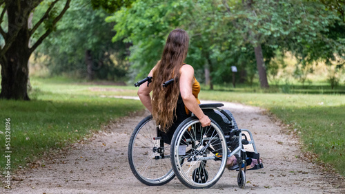 Young woman with long hair, moving in a wheelchair, in profile, in the middle of an alley lined with tall green trees