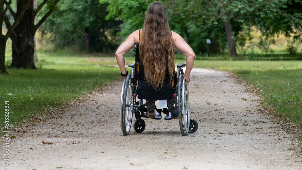 Young woman with long hair, moving in a wheelchair, from behind, in the middle of an alley lined with tall green trees Young woman with long hair, moving in a wheelchair, from behind, in the middle of an alley lined with tall green trees