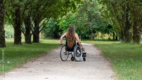 Young woman with long hair, moving in a wheelchair, from behind, in the middle of an alley lined with tall green trees