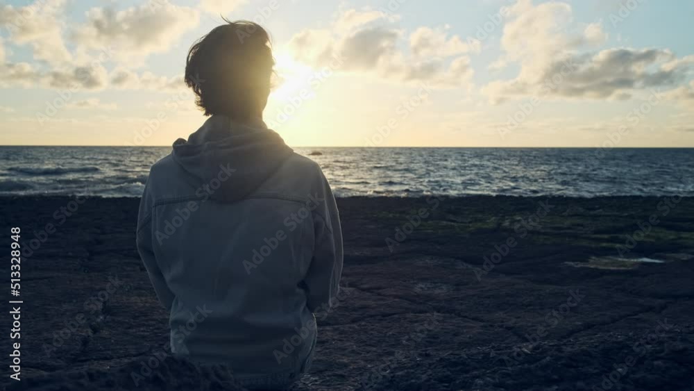 Human looks to the sun over horizon while sunrise. Man on a beach is looking distance during summer sunset. Person contemplates the beauty of nature. Freedom. Slow motion. Cinematic dramatic look. Human looks to the sun over horizon while sunrise. Man on a beach is looking distance during summer sunset. Person contemplates the beauty of nature. Freedom. Slow motion. Cinematic dramatic look.