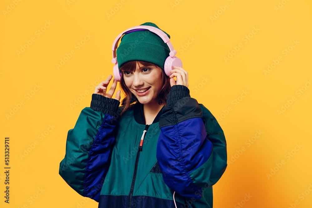a happy, joyful woman in a green jacket and hat enjoys music standing in pink headphones on a yellow background holding them with her hands and looking away with a pleasant smile on her face