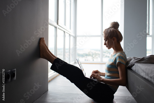 Blonde girl with hair bun working home, typing on laptop, sitting on the floor.