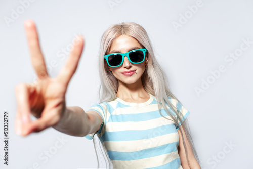 Portrait of cute blonde girl in striped shirt showing peace gesture on white.