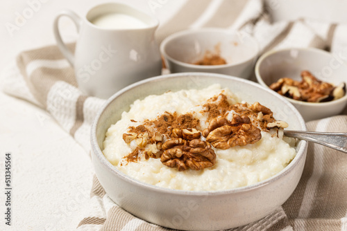 Creamy rice pudding whith milk, cinnamon powder and walnuts in white bowl with spoon on white table. Rice porridge with milk and topping for breakfast