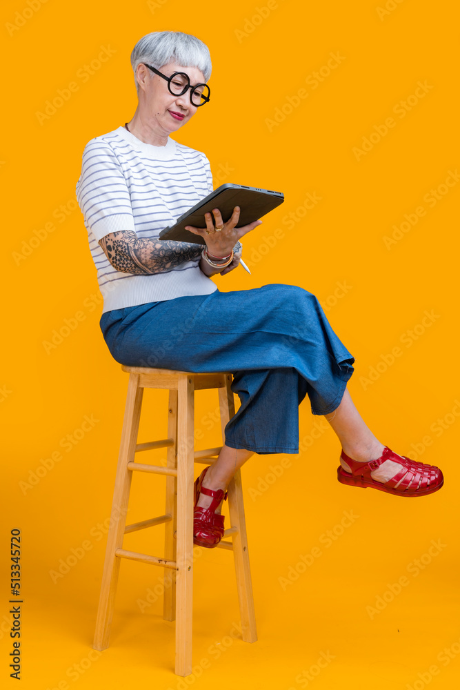 smiling retired old senior asian female with tattoo relax on comfy stool use modern digital touchpad tablet device read ebook from screen scroll social media web pages isolate yellow background