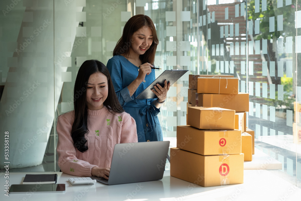 Young women teamwork partners packing order into box together using touchpad to do delivery to client. two asian female colleagues with tablet checking goods number and prepared cardboards in office. Young women teamwork partners packing order into box together using touchpad to do delivery to client. two asian female colleagues with tablet checking goods number and prepared cardboards in office.