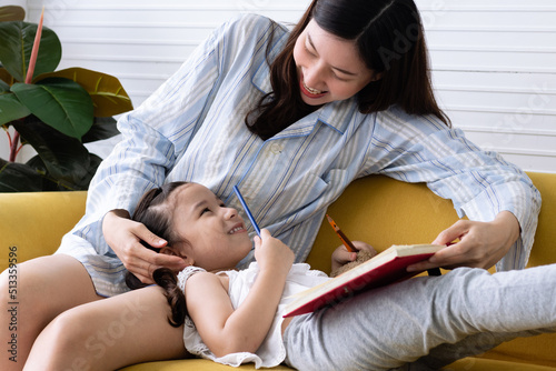 Daughter laying on mom's lap, reading and writing together on couch at home. Happy family and homeschool concept. Asian woman and Caucasian girl in pajamas playing at home.
