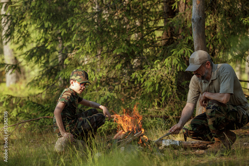 Father with son in military clothes is making campfire in the woods. The concept of adventure, travel, tourism, camping, survival and evacuation.
