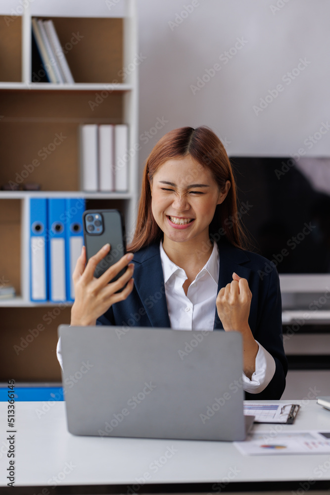 Happy Excited Asian young entrepreneur business woman using phone and laptop sitting on a desk officer in the day at the office Happy Excited Asian young entrepreneur business woman using phone and laptop sitting on a desk officer in the day at the office