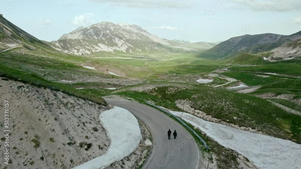 Two person are walking on tarmac curvy mountains road. On a beautiful sunny day. In the Italian mountains of Gran Sasso in the Abruzzo region. Two person are walking on tarmac curvy mountains road. On a beautiful sunny day. In the Italian mountains of Gran Sasso in the Abruzzo region.