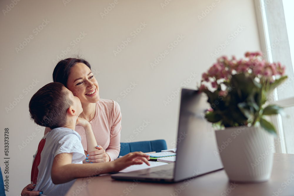 Little cute son kissing his mother in cheek while studying online at laptop at home.