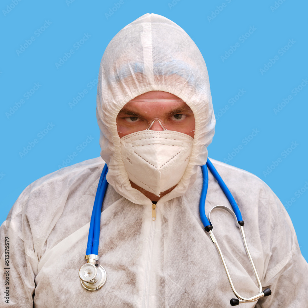 Man doctor medic in a protective suit uniform with goggles and face mask on a studio blue background. Paramedic in white antiviral protective clothing wearing an N95 respirator and safety glasses Man doctor medic in a protective suit uniform with goggles and face mask on a studio blue background. Paramedic in white antiviral protective clothing wearing an N95 respirator and safety glasses