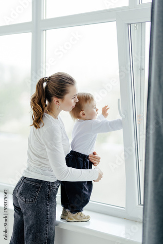 Portrait of a smiling young cute mother and her son playing near the window in a room. Sunny day outside. True mother love