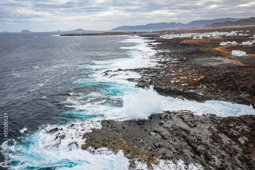 aerial view of ocean waves crashing on the rocks with mountains and village in background 