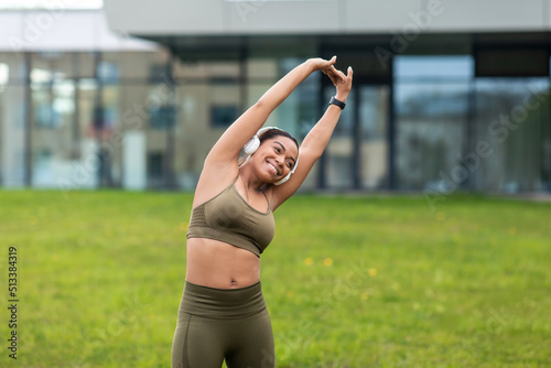 Happy young black woman listening to music in headphones, stretching her arm, doing sports at city park, copy space
