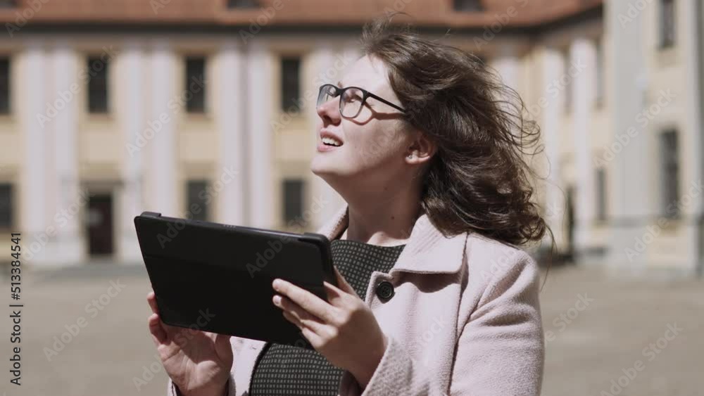 Young pretty woman in glasses is looking for information about sights in tablet. Lady in courtyard of medieval castle, museum, clicks on gadget screen, smiles. Sunny day, wind blows on curly hair.