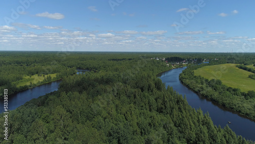 Narrow curved river surrounded by deciduous trees on the banks on a sunny spring day. Shot. Aerial view of a quiet village with small houses on blue cloudy sky background.