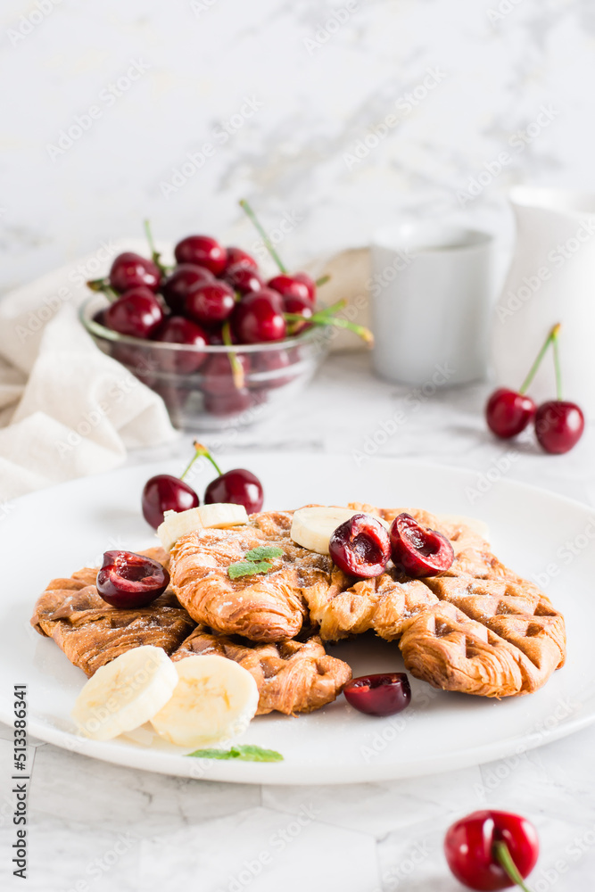 Appetizing croffles with cherries and bananas on a plate on the table. Street food. Vertical view Appetizing croffles with cherries and bananas on a plate on the table. Street food. Vertical view