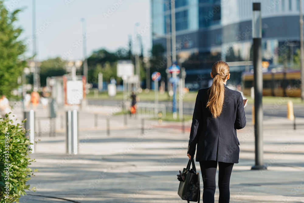 A female realtor in a blazer is walking with a laptop between skyscrapers.