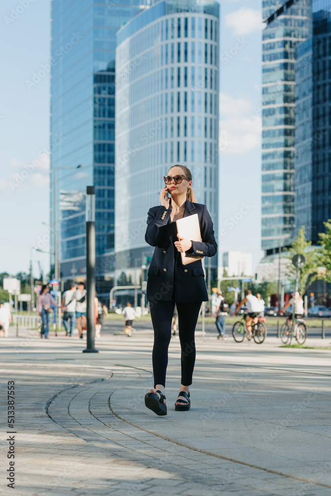 A female realtor in a navy blue blazer is walking with a laptop and having a conversation on a cell phone in the financial district. A businesswoman in a cluster of tall buildings.