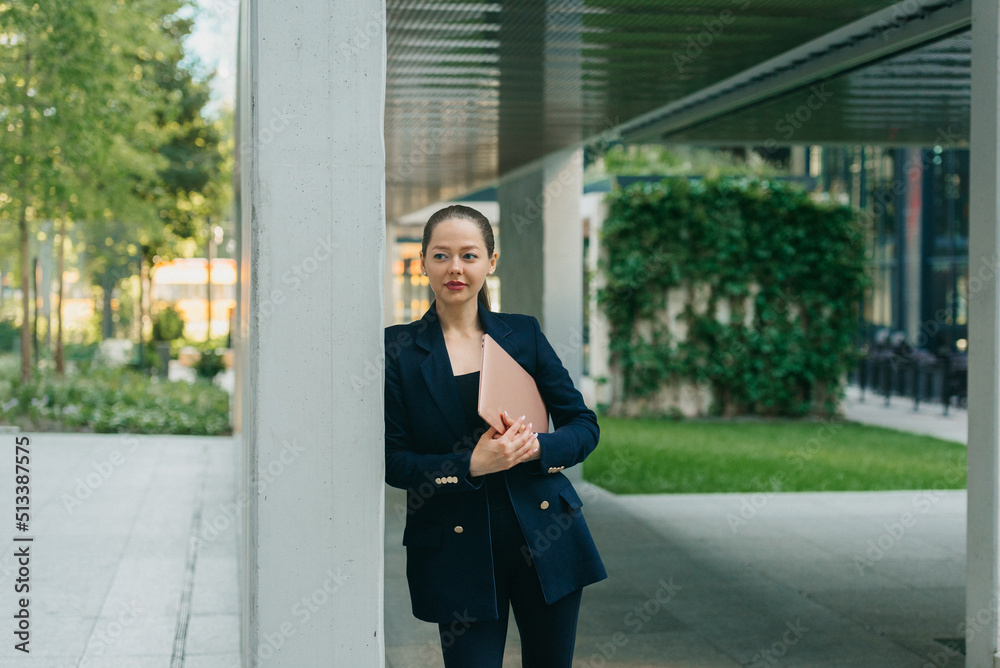 A female realtor in a blazer is holding a computer while leaning against the wall between the office buildings in the financial district. A pretty businesswoman in the contemporary urban space. A female realtor in a blazer is holding a computer while leaning against the wall between the office buildings in the financial district. A pretty businesswoman in the contemporary urban space.