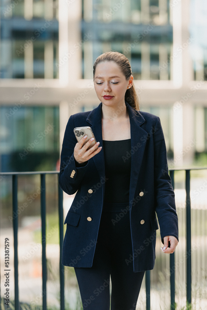 Fototapeta premium A female realtor in a blazer is checking the news on her smartphone in a modern urban space in the financial district. A businesswoman in the contemporary garden in a cluster of tall buildings.