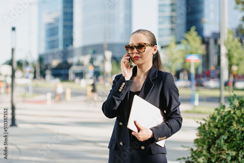 A female realtor in a blue blazer is holding a laptop between the office buildings in the financial district. A businesswoman is having a conversation on her smartphone in a cluster of tall buildings.