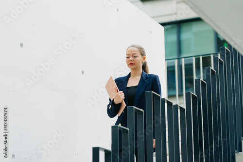 A female realtor in a blazer is descending the stairs with a computer between the office buildings in the financial district. A pretty businesswoman in the contemporary urban space.