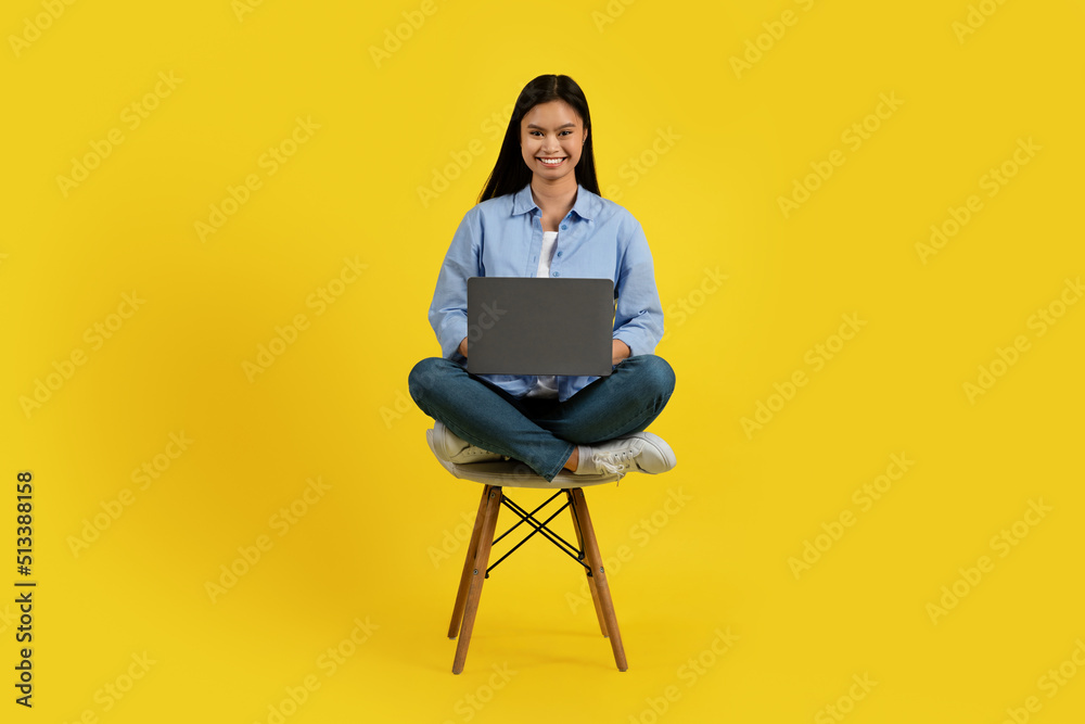 Cheerful young chinese lady student sitting on chair and working on laptop, isolated on yellow background Cheerful young chinese lady student sitting on chair and working on laptop, isolated on yellow background