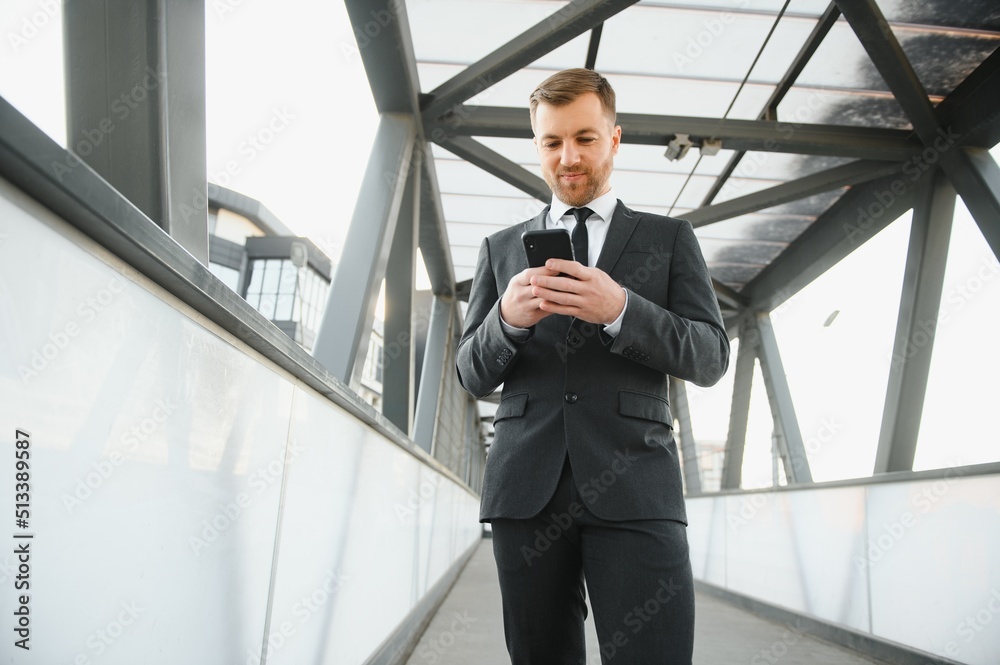 Happy smiling businessman wearing black suit and using modern smartphone near office at early morning, successful employer to make a deal while standing near skyscraper office at night, flare light Happy smiling businessman wearing black suit and using modern smartphone near office at early morning, successful employer to make a deal while standing near skyscraper office at night, flare light