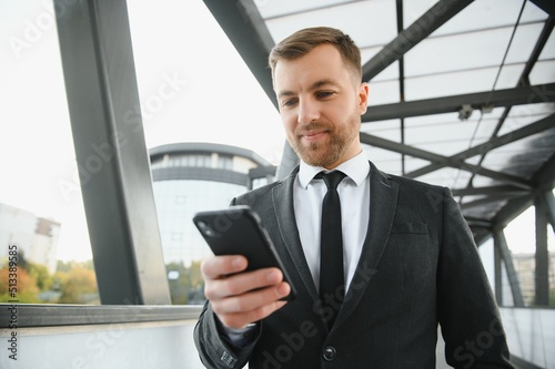 Happy smiling businessman wearing black suit and using modern smartphone near office at early morning, successful employer to make a deal while standing near skyscraper office at night, flare light