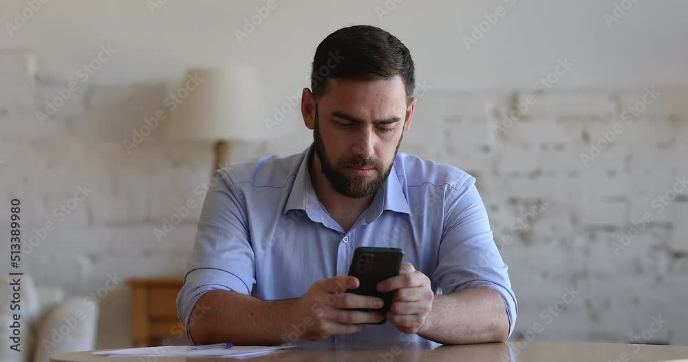 Focused millennial freelance employee man typing on mobile phone at home table, chatting online, reading text message with good news, getting happy, smiling, looking away