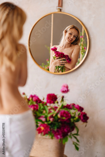 a blonde woman with a bare back and a bouquet of peonies looks at in the mirror.