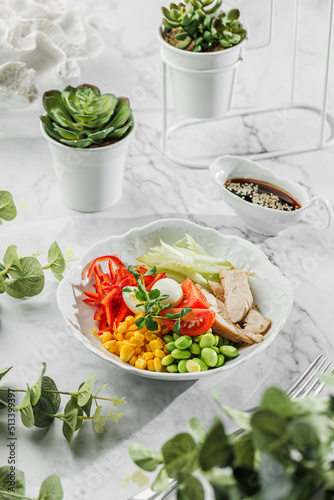 Poke bowl , buddha boul with boled chicken ,boiled eggs, avocado, cherry, corn, cabbage and greens at the white plate on light marble background. Healthy sea food, hard light, restaurant decor photo