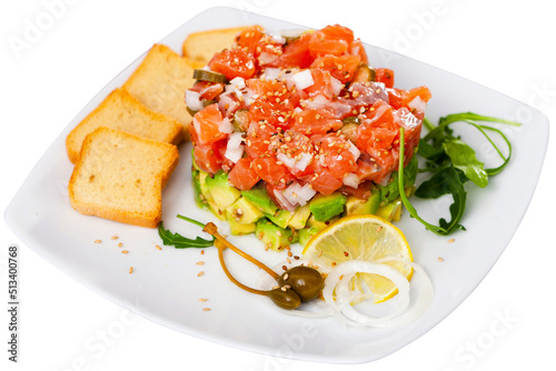 Raw salmon tartare with diced avocado sprinkled with sesame seeds served with crispy toasts, arugula, lemon and pickled capers on white plate. Isolated over white background