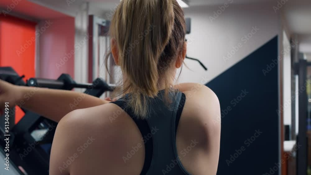 Rear view of a woman in the gym stretching her shoulder joints before a workout. A woman is doing a warm-up before exercising in the gym. The concept of exercising in the gym. Rear view of a woman in the gym stretching her shoulder joints before a workout. A woman is doing a warm-up before exercising in the gym. The concept of exercising in the gym.
