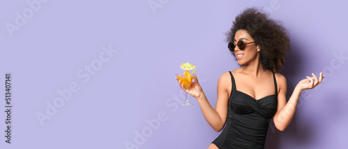 Beautiful African-American woman in bikini and with cocktail on lilac background with space for text