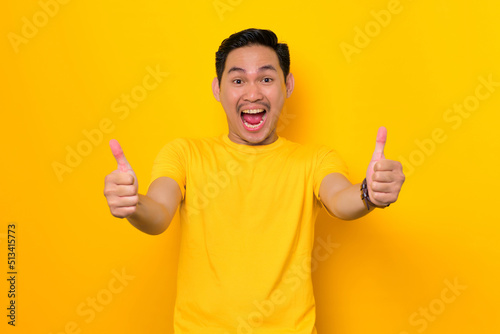 Cheerful young Asian man in casual t-shirt showing thumb up and gesturing sign of approval isolated on yellow background. People lifestyle concept