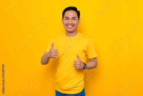 Smiling young Asian man in casual t-shirt showing thumb up and gesturing sign of approval isolated on yellow background. People lifestyle concept