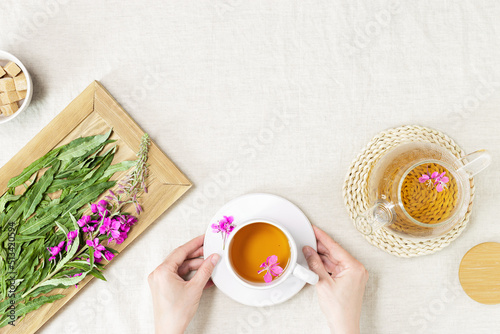 Woman hands holding Herbal tea from kipreya leaves in cups on fabric table background, fireweed green leaf and flower on wooden tray. Flavored herbal tea from natural wild plants, healing beverage