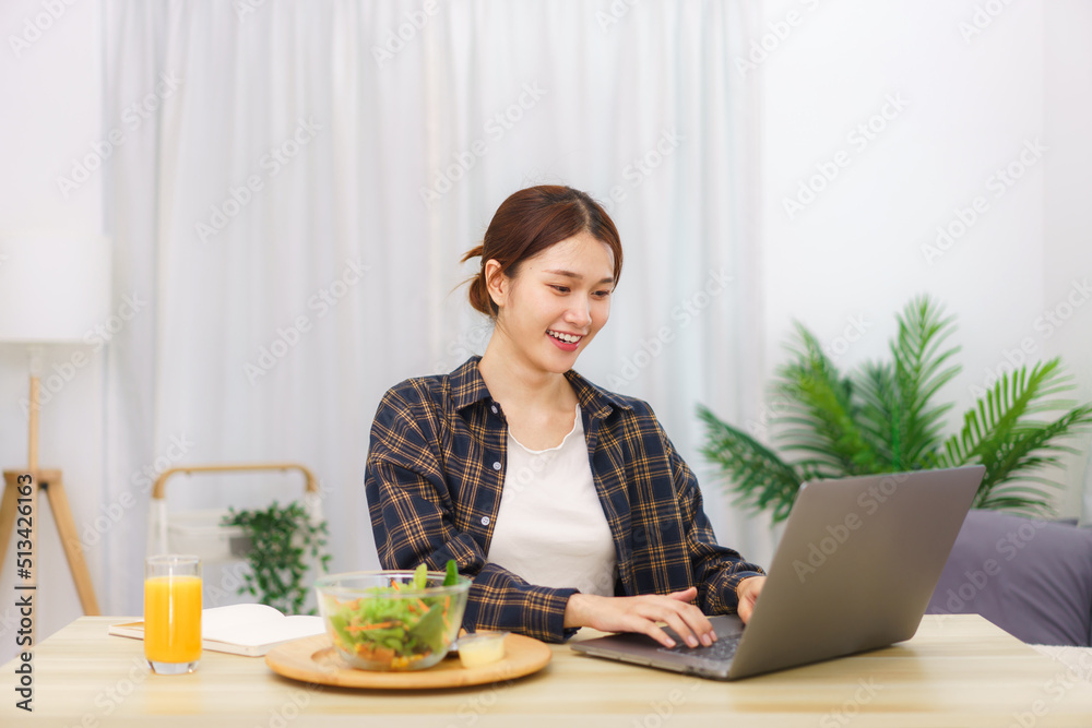 Lifestyle in living room concept, Young Asian woman eating vegetable salad and working on laptop Lifestyle in living room concept, Young Asian woman eating vegetable salad and working on laptop