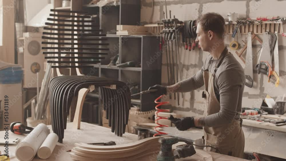 Medium slowmo of young carpenter dying designer authentic chair in black color, finishing his project in workshop