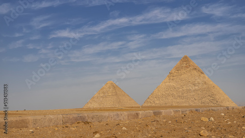 Two great pyramids of Cheops and Chephren on a background of blue sky and clouds. In the foreground is the sandy-rocky soil of the Giza plateau  a road curb. Egypt